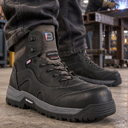 Black US Boot Iron Master boots with gray midsoles, with a welder working at a steel bench in the background.
