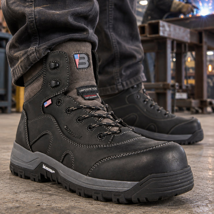 Black US Boot Iron Master boots with gray midsoles, with a welder working at a steel bench in the background.