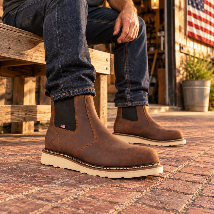 Seated craftsman wearing brown US Boot James Chelsea boots with cream wedge soles outside a workshop in warm evening light.