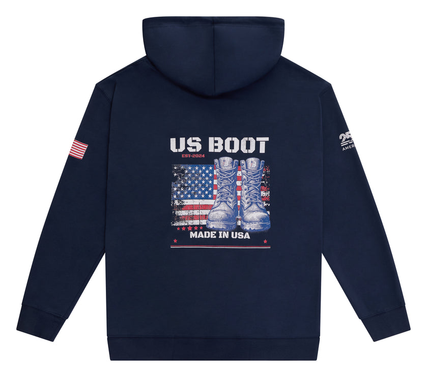 A navy blue hooded sweatshirt shown from the back, featuring a large graphic print of combat boots over an American flag with the text 'US BOOT' and 'MADE IN USA'. A small American flag graphic is on the right sleeve, and a white graphic is on the left sleeve.